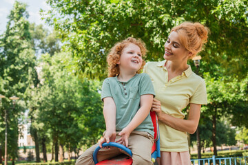 happy redhead woman smiling near son riding seesaw on playground.