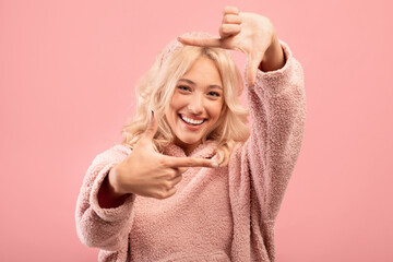 Capturing moments. Happy young lady making picture frame with fingers, looking at camera and smiling, pink background