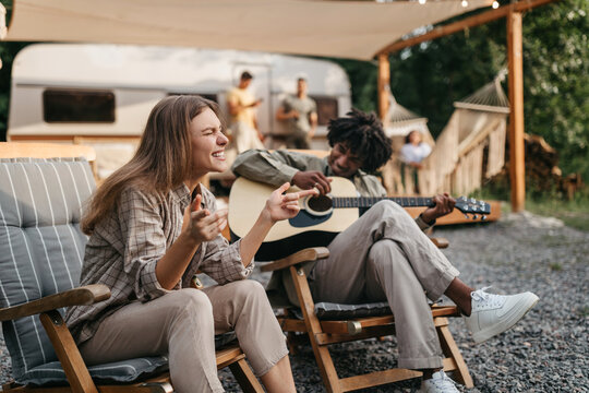 Happy black guy playing guitar, his female friend singing, sitting in lounge chairs near RV, camping on summer vacation