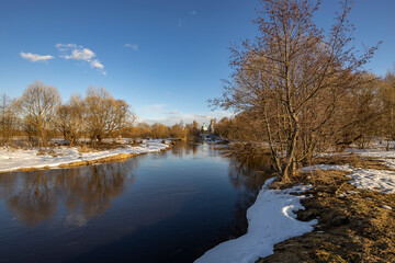 A picturesque landscape, early spring, a river with snow-covered banks, dry grass and bushes. March sunny day by the river. Church in the background. The first thaws, the snow is melting.