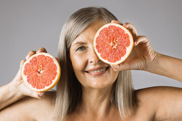 Smiling caucasian woman with naturally aged skin posing over grey background with slices of sweet grapefruit in hands. Concept of people, natural cosmetic and beauty treatment.