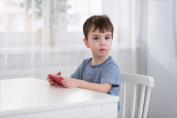 A cute little boy is sitting at the table and playing on the phone. Gadgets and kids concept