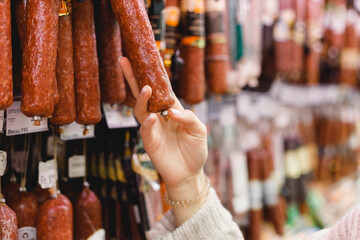 Woman chooses dried smoked sausage hanging in refrigerator in butcher's shop