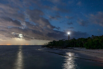 Near Fort Myers, Florida, the rising moon shines on San Carlos Bay as the Ybel Lighthouse shines on the beach at the tip of Sanibel Island while stars twinkle in the cloudy night sky. - 495518374