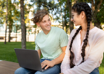 Excited students lady and guy studying together in park, preparing for exam and using laptop, sitting in park