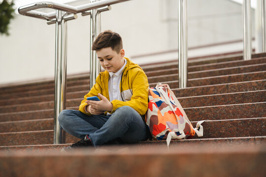 Cute schoolboy in a yellow jacket sits on the stairs and uses a smartphone