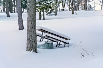 Snow covered picnic table leaned against a tree and fire pit in campground at Interlochen State Park, Michigan