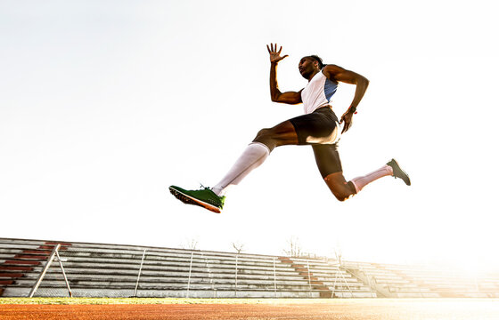 Professional african american male track and field athlete during obstacle race - Young athlete performing explosive long jump during training on race track in athletics stadium - Freedom concept