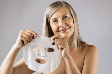 Attractive mature lady with bare shoulders holding in hands cotton face mask over grey background. Portrait of happy woman using cosmetic for facial treatment.