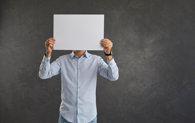 Man holding blank white sheet of paper in front of his face showing copy space on camera. Unknown...