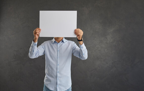 Man holding blank white sheet of paper in front of his face showing copy space on camera. Unknown and unrecognizable young man in shirt standing on gray background hides his face behind sheet of paper