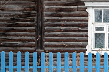 White window on the wall of a log house. A blue fence in front of a wall of a log house. Old logs of brown color on the wall of a house with one window.A blue fence in front of a house.