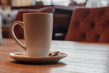 Tall white coffee mug on a saucer with a teaspoon on a wooden table in a cafe. Hot invigorating drinks for breakfast. The concept of a cozy breakfast. Space for text.