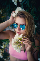 Attractive young woman in a pink swimsuit, sunglasses on her face stands by a plant with white flowers