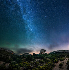 Panoramic view of the Milky Way and the Andromeda galaxy over a rock arch shaped like a turtle. 