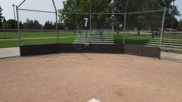 Pan out of ball field number 7 from home plate to second base. Foot prints in the sand. Bases in the field. Empty bleachers waiting. Trees in quiet park. Roof over dugouts. 