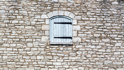 Wall of an old stable building made of stone blocks with a window that has a wooden shutter with large black metal decorative hinges