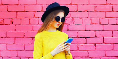 Portrait of young woman with smartphone wearing black round hat, yellow sweater on pink background