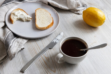 A cup of coffee, two pieces of sugar, a lemon, a piece of bread with curd cheese spread on it on a...