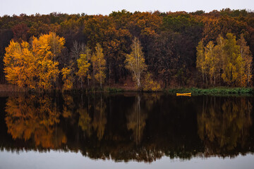 Yellow small boat near the forest with golden autumn trees, reflections in water