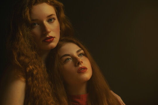 Studio portrait of two beautiful redhead freckled women with red lips makeup, long hair posing in darkness and warm light