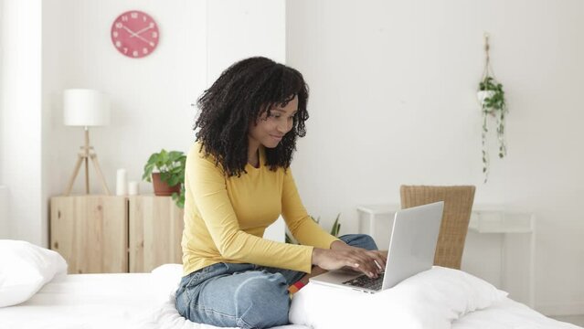 Happy young african american woman working on laptop in bed. Brazilian female using computer while relaxing in bedroom - Technology concept - High quality 4k footage
