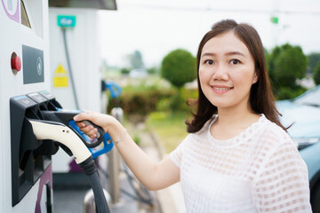 Happy Asian woman holding a DC - CCS type 2 EV charging connector at EV charging station.