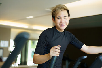 Active Asian young man making a cardio exercise on the elliptical exercise machine in the gym. Fat...