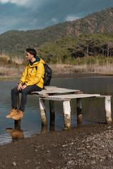 Alone young man sitting on old small dock. He is watching beautiful view. Cloudy sky background. Vertical photo.	