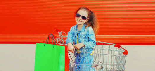 Portrait of happy smiling little girl child with trolley cart and shopping bags wearing denim clothes on city street