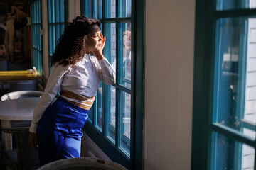 Fototapeta na wymiar Happy curly brunette girl smiling in cafe. Young woman enyoing sun in restaurant at window. white blouse. Sun in city. Fashionable asian girl with frizzly hair. Sun light in window