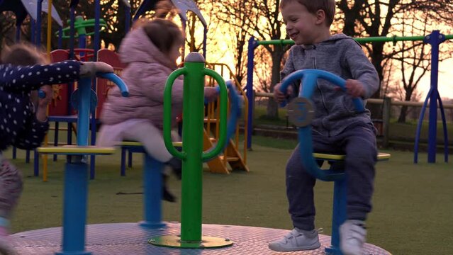Three little Happy Children having fun spinning on merry-go-round. Smiling Siblings friends playing outside. emotional kids rides on carousel at playground nursery in park.Happy family, childhood