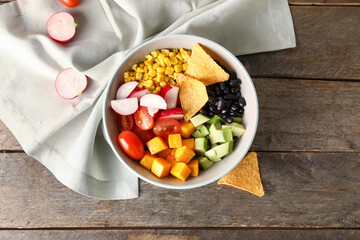 Bowl of tasty Mexican vegetable salad with black beans, radish and nachos on wooden background