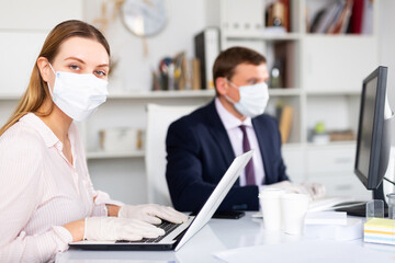 Focused young woman in medical face mask and latex gloves working on laptop in office with male colleague. Concept of prevention of viral infection and social distancing