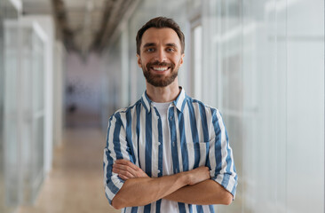 Obraz premium Portrait of handsome smiling businessman with arms crossed looking at camera standing in modern office 