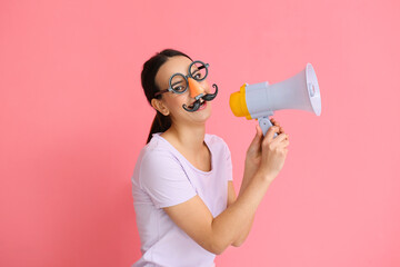 Young woman with funny disguise shouting into megaphone on pink background. April Fools Day celebration