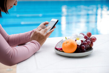 Hand of an Asian woman in a swimsuit sitting by the hotel pool Sliding mobile phone for online shopping with fruit tray