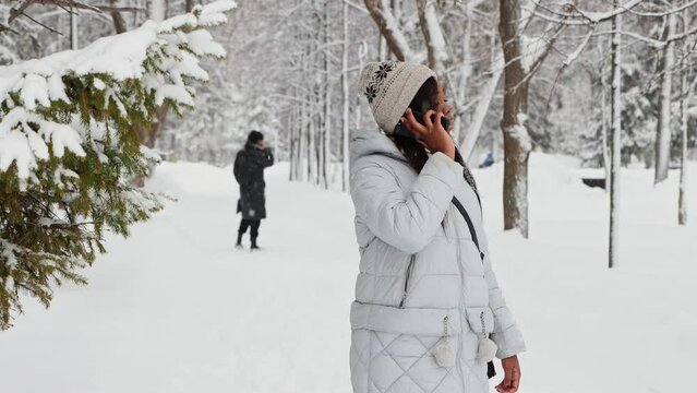 Young black woman talks on her phone in winter forest and finds her friend behind her