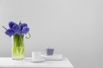Vase with hyacinth flowers, cup and candle on table near light wall