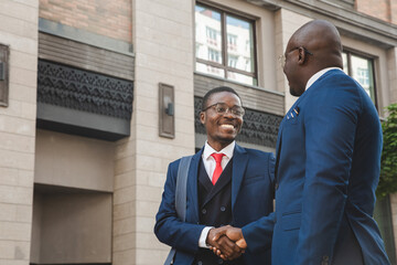 Two black african american businessmen in suits and glasses shake hands while meeting outdoors