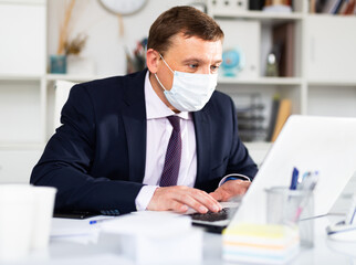 Portrait of confident manager wearing medical mask working on laptop in office. New life reality during coronavirus pandemic..