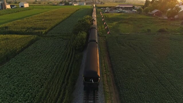An Aerial From Behind the Train View of A Steam Passenger Train Passing Thru Farmlands During the Golden Hour on a Summer Day
