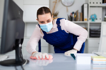 Fototapeta na wymiar Woman professional cleaner in protective medical mask cleaning desk with detergents in office