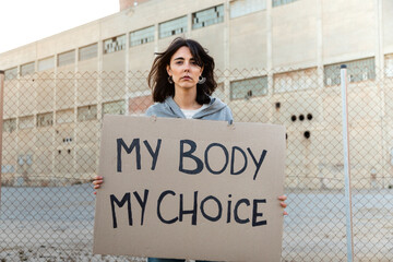 Young caucasian woman looking at camera with serious expression holding a cardboard sign: My body my choice.