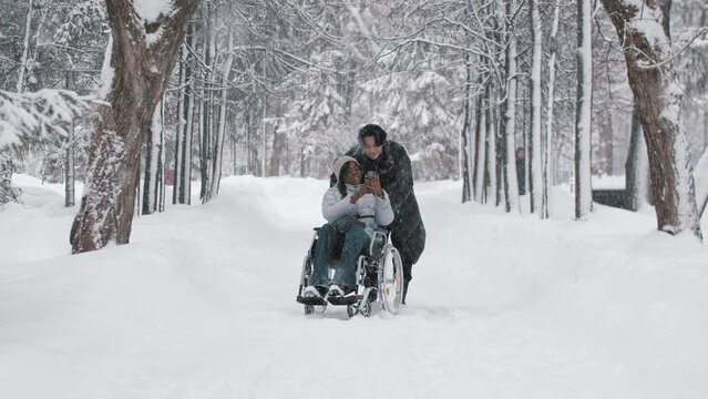 Young couple of asian guy and his black girlfriend in the wheelchair taking a selfie in winter forest