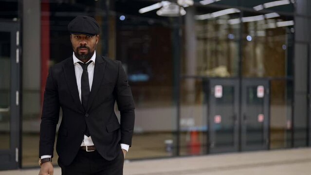 serious afro-american man dressed in black suit and cap is strolling in mall, moving to camera