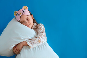 Portrait of a positive cheerful child in an eye mask, hugging her big soft pillow on a blue...