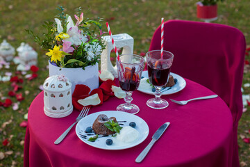Romantic table for two outdoors with a red tablecloth and covers on chairs, with desserts, mulled wine, a bouquet of flowers, candles and rose petals. The concept of marriage proposal.
