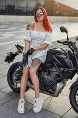 Fototapeta na wymiar Redhead woman biker posing on black motorcycle outdoors
