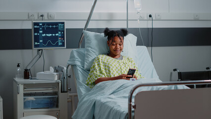 Patient with disease using smartphone to text messages and browse internet while laying in hospital ward bed. Young woman holding mobile phone to have fun while recovering from sickness